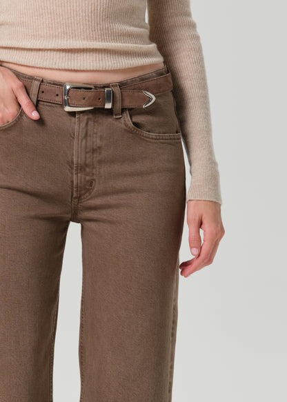 Close-up of a person wearing brown jeans with a matching brown belt featuring a silver-toned buckle, paired with a beige ribbed knit top against a grey background.