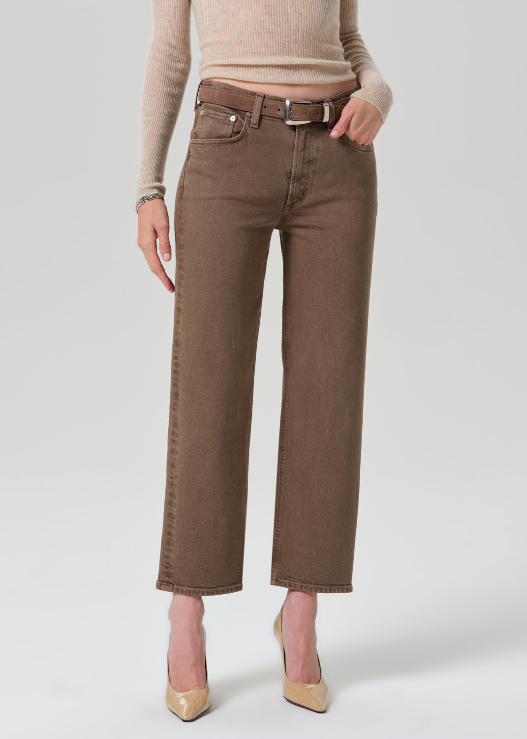 A model wearing brown cropped jeans with a matching brown belt, paired with a beige ribbed sweater and beige heels, shown from the waist down on a grey background.