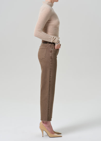 A side-profile view of a model wearing brown slim-fit jeans, a beige ribbed turtleneck sweater, and beige high-heeled pumps against a grey background.