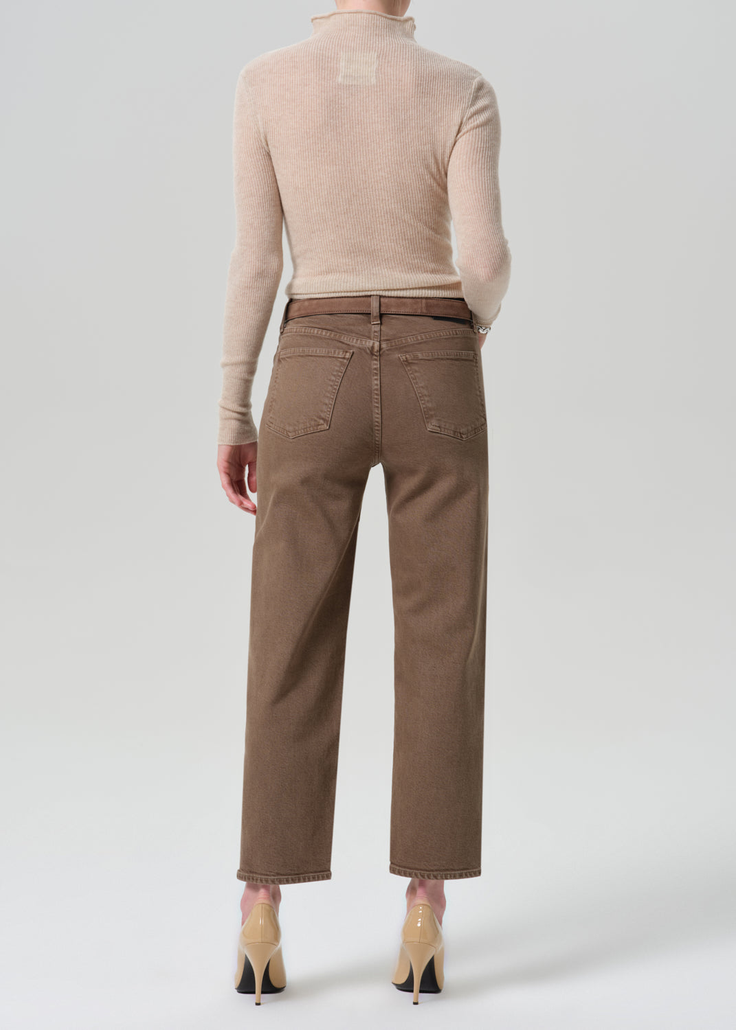 A model shown from the back wearing a light beige mock-neck knit top and brown cropped jeans, paired with beige high heels, against a grey background.