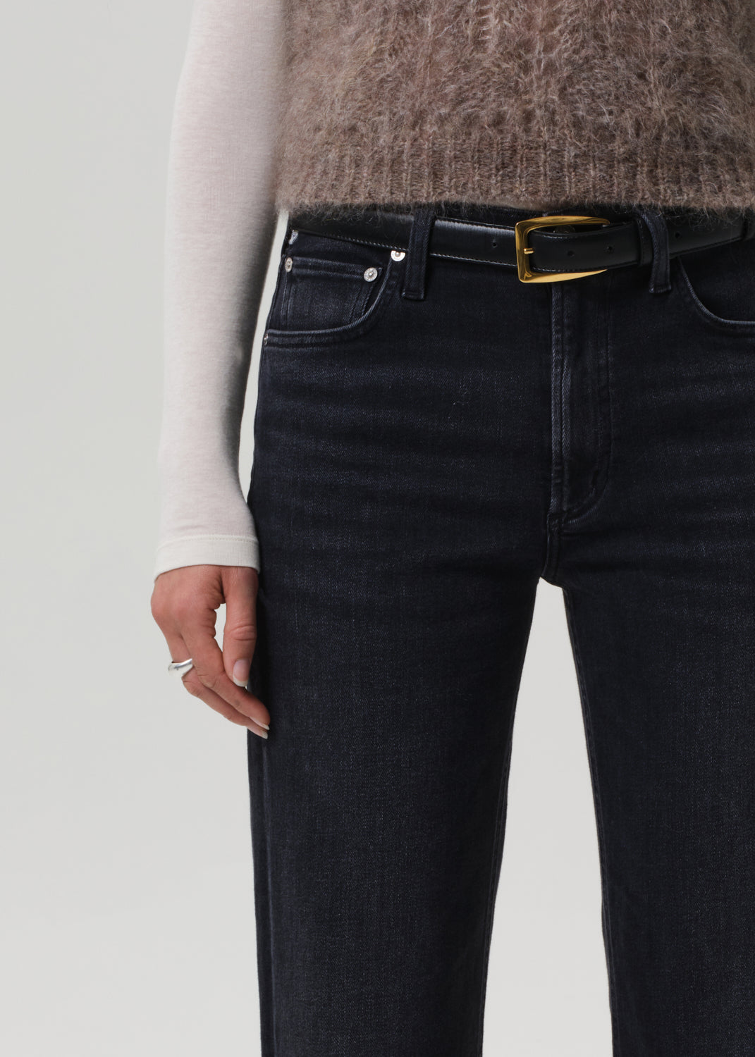 Person wearing a brown knitted sweater, white turtleneck, black jeans, and black boots on a light gray background