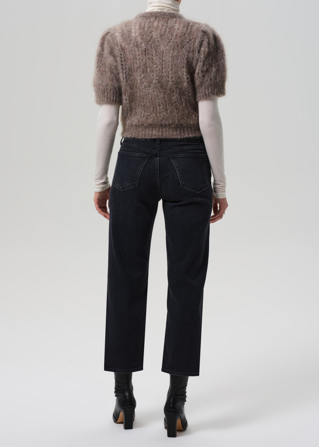 Person wearing a brown knitted sweater, white turtleneck, black jeans, and black boots on a light gray background