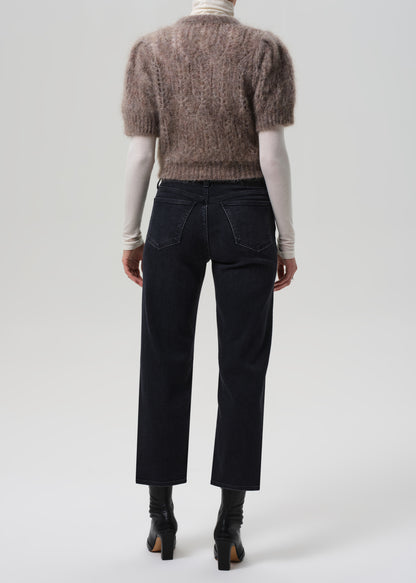 Person wearing a brown knitted sweater, white turtleneck, black jeans, and black boots on a light gray background