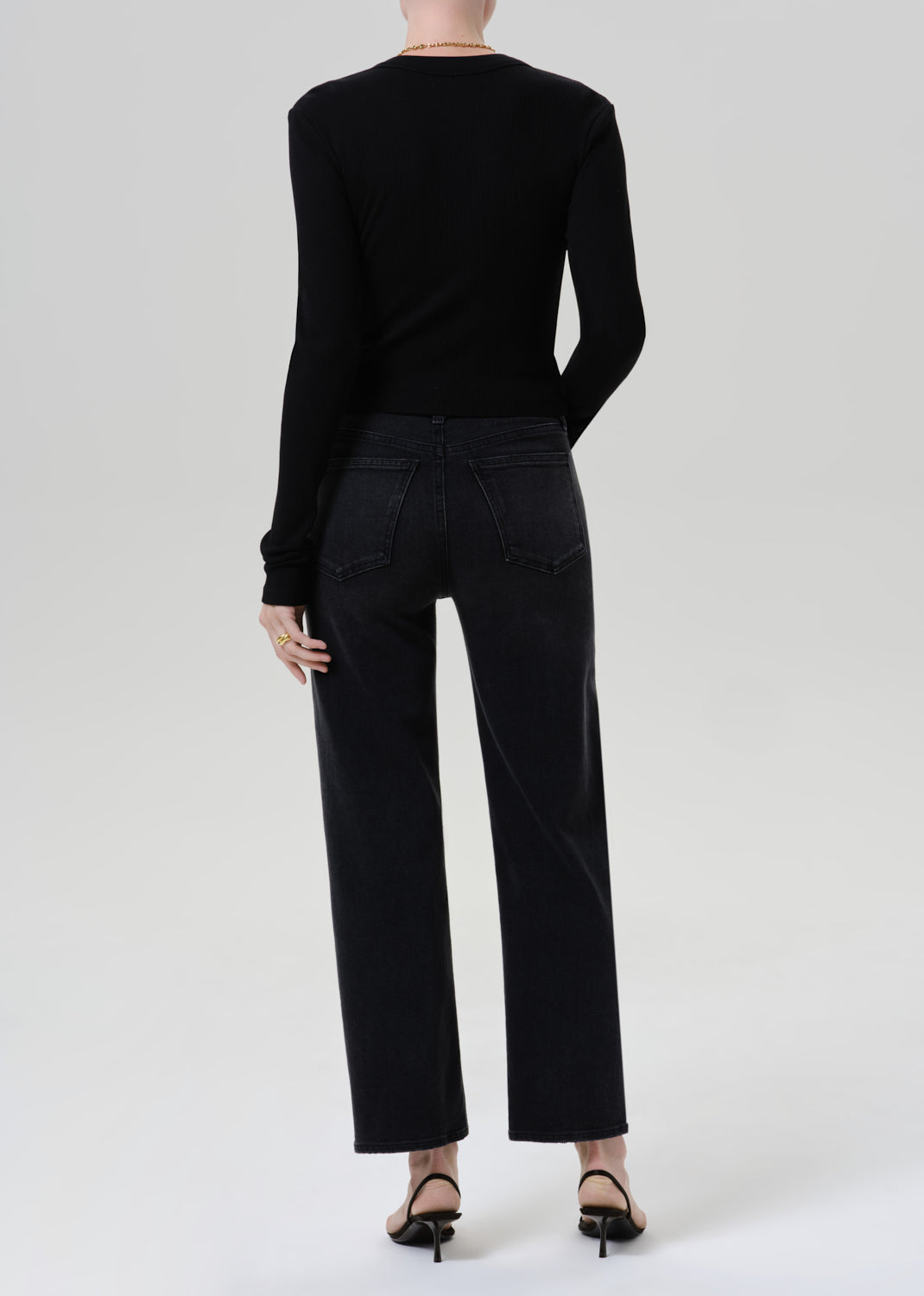 Person wearing a black long sleeve henley top with black straight leg jeans viewed from the back on a grey background