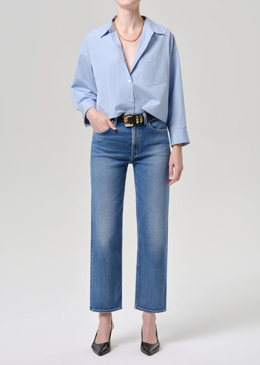 A model wearing a light blue button-down shirt and blue cropped jeans with a black belt, paired with black pointed-toe heels, against a grey background.