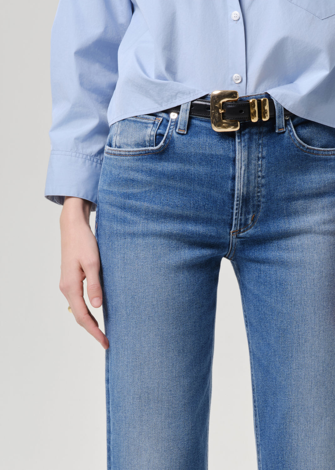Close-up of a person wearing blue jeans with a black belt featuring a prominent gold buckle, paired with a light blue button-up shirt.