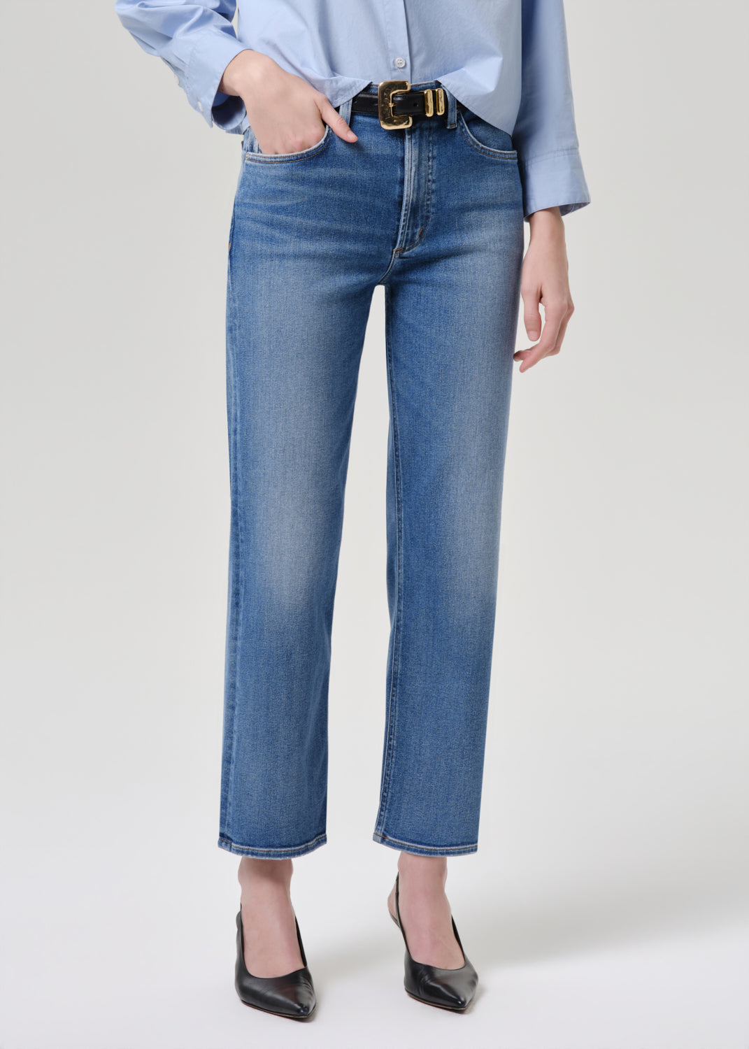 A model wearing a pair of medium-wash blue denim jeans with a straight-leg fit, styled with a black belt, a light blue shirt, and black pointed-toe heels against a grey background.