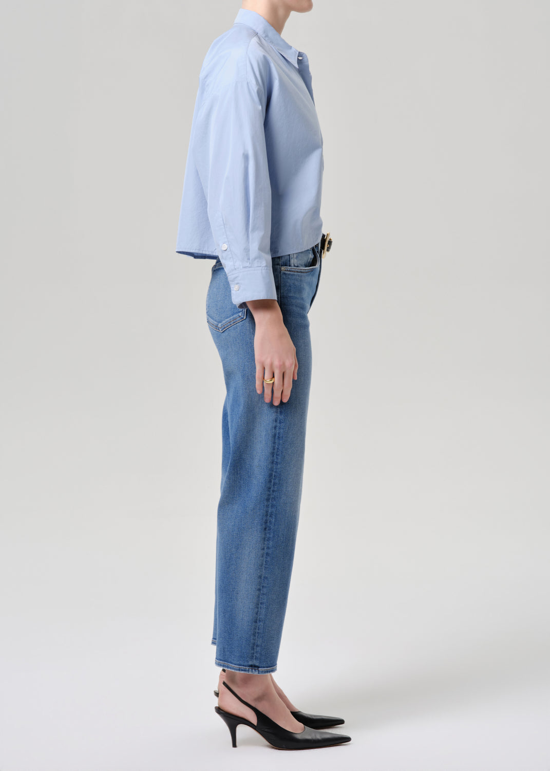 A side-profile studio shot of a model wearing a light blue long-sleeve button-up shirt tucked into light-wash blue jeans, paired with black slingback heels, against a grey background.