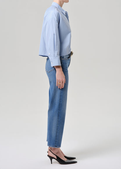 A side-profile studio shot of a model wearing a light blue long-sleeve button-up shirt tucked into light-wash blue jeans, paired with black slingback heels, against a grey background.
