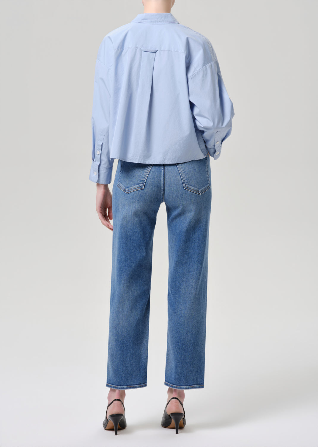 A model shown from the back wearing a light blue cropped button-down shirt and medium-wash blue jeans, paired with black high-heeled sandals, against a grey background.