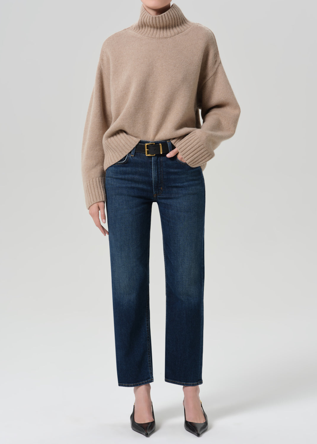 Person wearing a beige turtleneck sweater and dark blue pants on a grey background