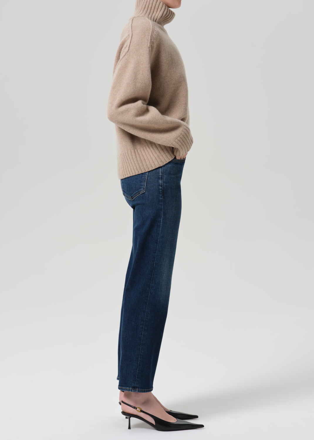 Person wearing a beige turtleneck sweater and dark blue pants on a grey background