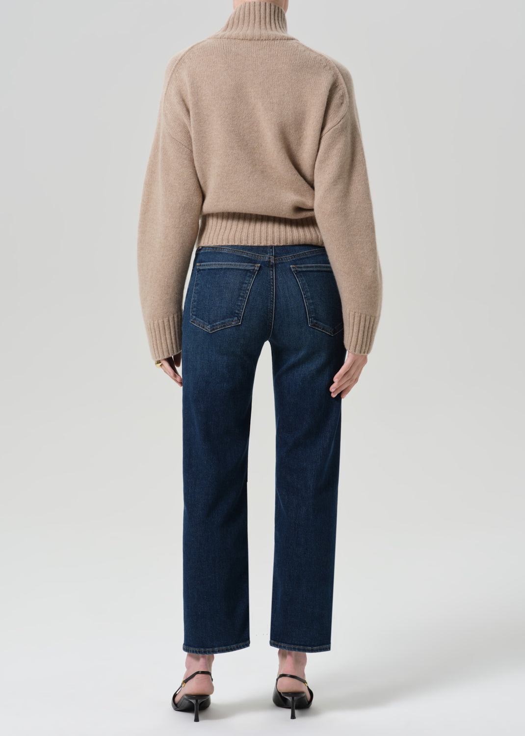 Person wearing a beige turtleneck sweater and dark blue pants on a grey background