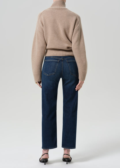 Person wearing a beige turtleneck sweater and dark blue pants on a grey background