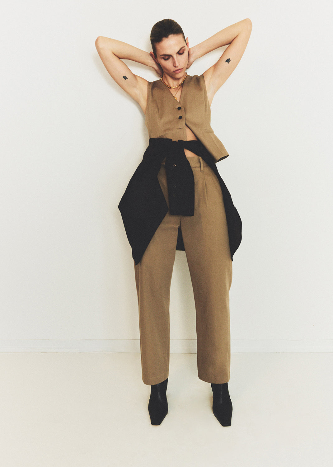 Woman wearing a beige sleeveless vest and pants with a black belt against a white background