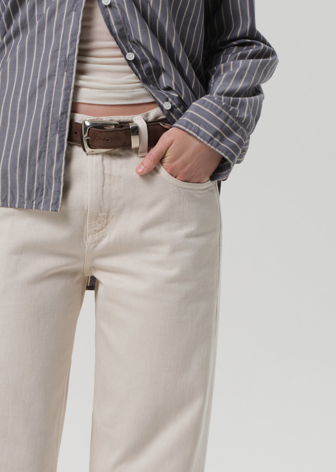 Person wearing a striped shirt, white top, beige pants, and brown belt on a plain background