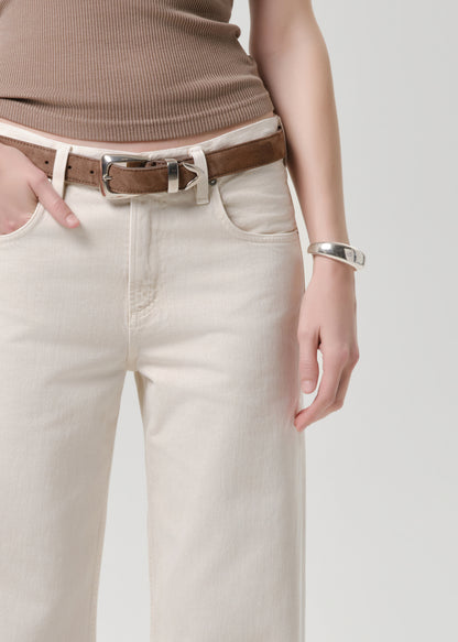 Close-up of a person wearing off-white straight-leg jeans, a brown leather belt with a silver buckle, a taupe ribbed top, and a silver cuff bracelet against a grey background.