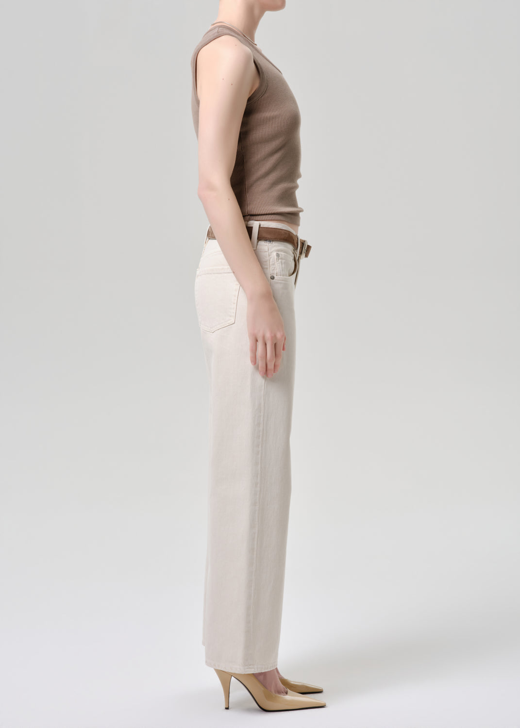 A side-profile studio shot of a model wearing a sleeveless taupe top, off-white wide-leg jeans, and beige heels against a grey background.