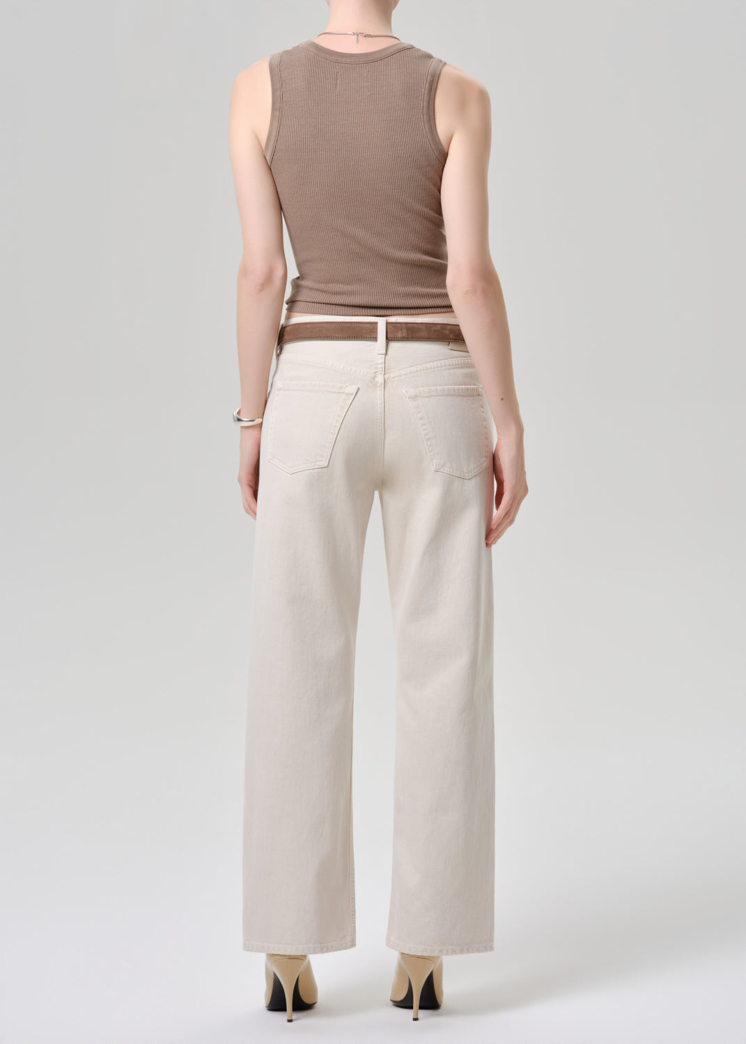 A model shown from the back wearing a sleeveless taupe-brown top, off-white wide-leg jeans, a brown belt, and beige heels against a grey background.