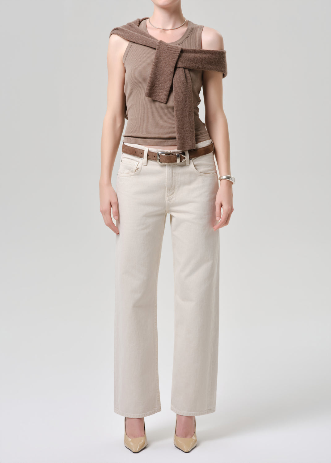 Woman wearing a brown top with a scarf, white pants, and beige shoes on a grey background