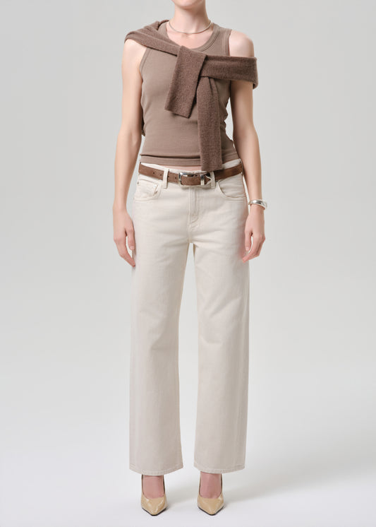 Woman wearing a brown top with a scarf, white pants, and beige shoes on a grey background