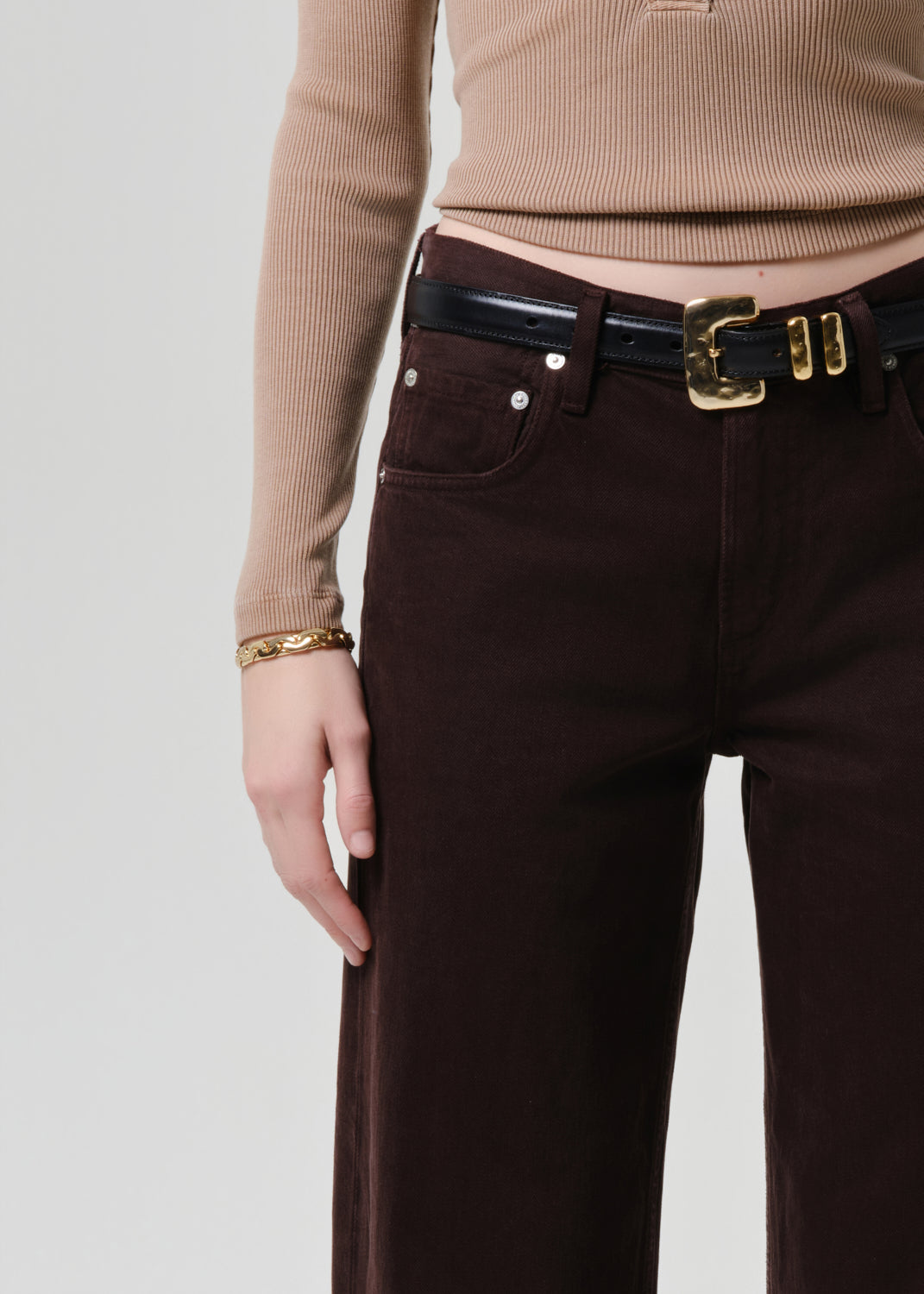 Close-up of a person wearing dark brown wide-leg trousers, a black belt with a gold buckle, a beige ribbed long-sleeve top, and a gold bracelet against a grey background.