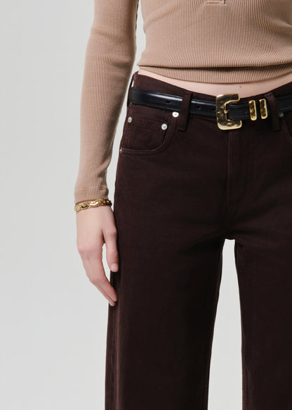 Close-up of a person wearing dark brown wide-leg trousers, a black belt with a gold buckle, a beige ribbed long-sleeve top, and a gold bracelet against a grey background.