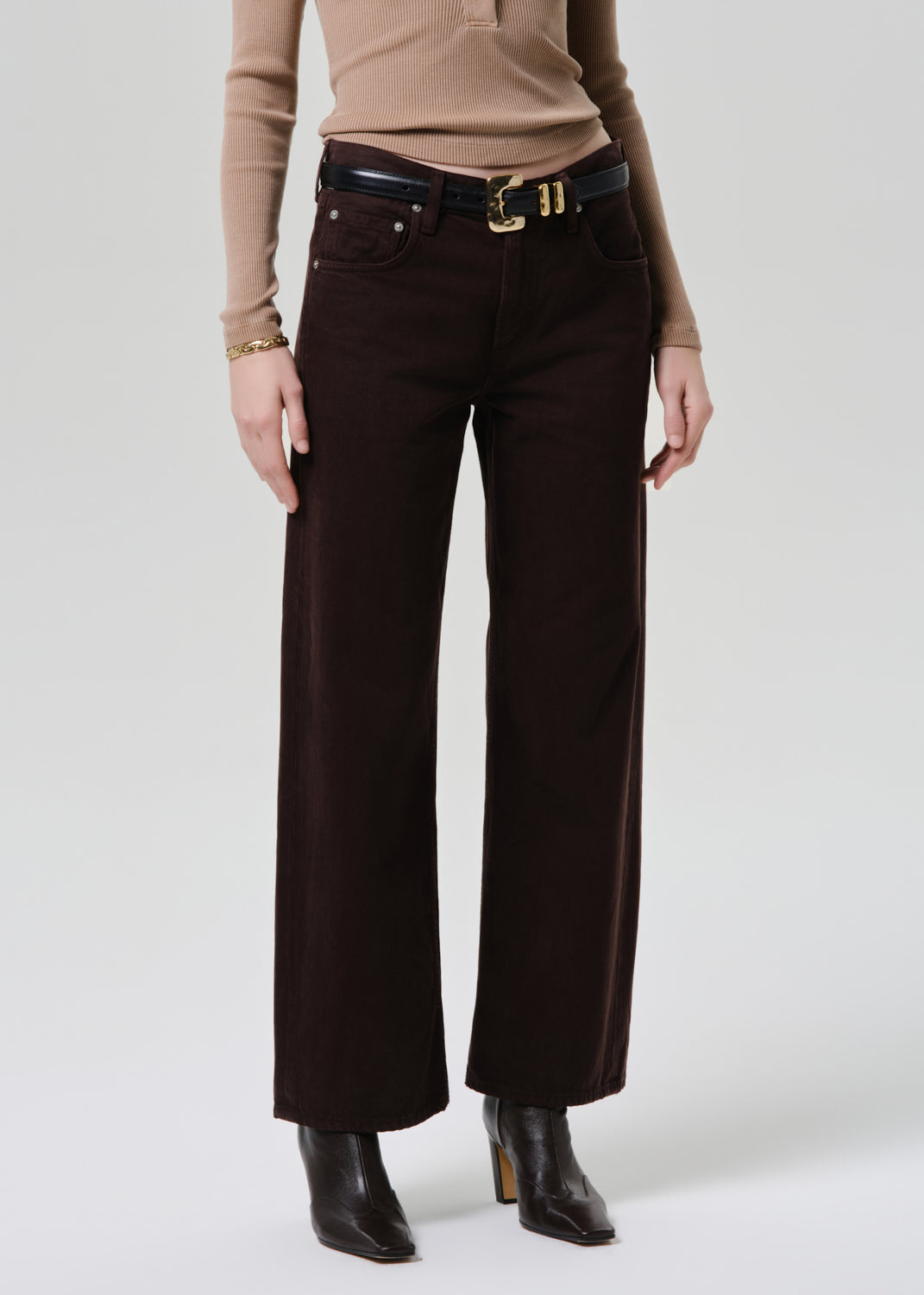 A model wearing dark brown wide-leg corduroy pants with a black leather belt featuring gold hardware, styled with a beige long-sleeve top and black ankle boots.