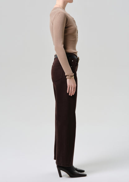 A side-profile studio shot of a model wearing dark brown wide-leg trousers, a beige ribbed long-sleeve top, a black belt, and black pointed-toe heels against a grey background.