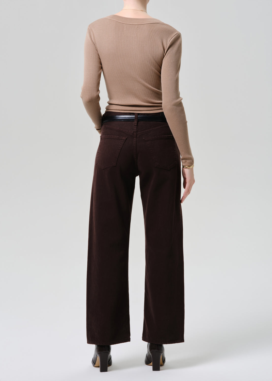 A model shown from the back wearing a beige long-sleeve top and dark brown wide-leg trousers, paired with a black belt and black heels, against a grey background.