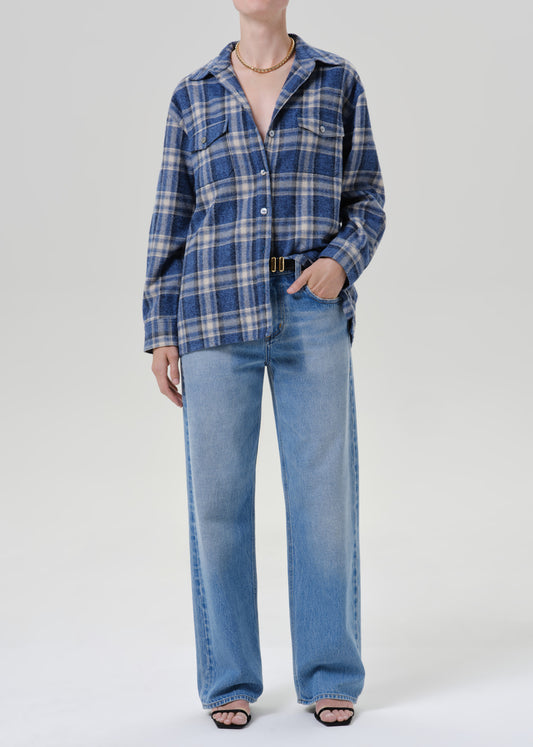 A model wearing a blue and white plaid button-up shirt with wide-leg light-wash blue jeans, styled with a black belt and black sandals against a grey background.