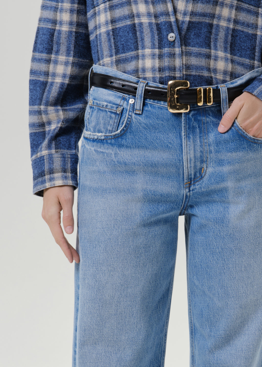 A close-up of a person wearing light blue jeans, a blue and white plaid flannel shirt, and a black leather belt with a prominent gold buckle.