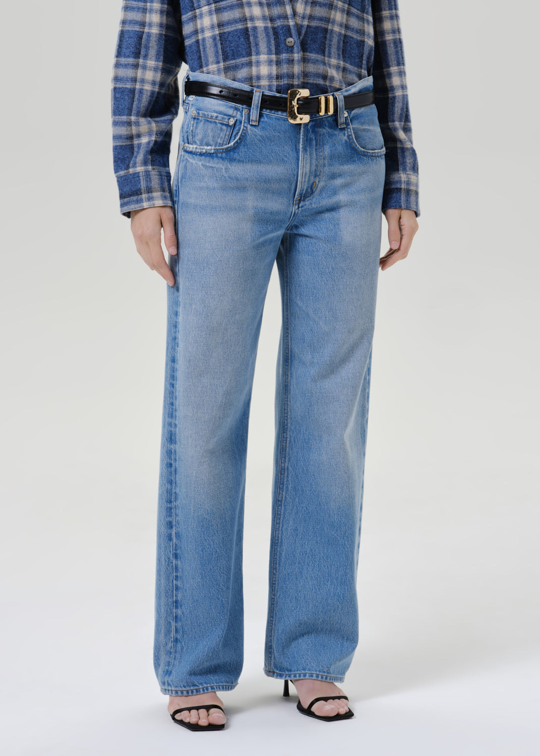 A model wearing a pair of light blue wide-leg denim jeans with a black belt featuring a gold buckle, styled with a blue and white plaid shirt and black heeled sandals against a grey background.