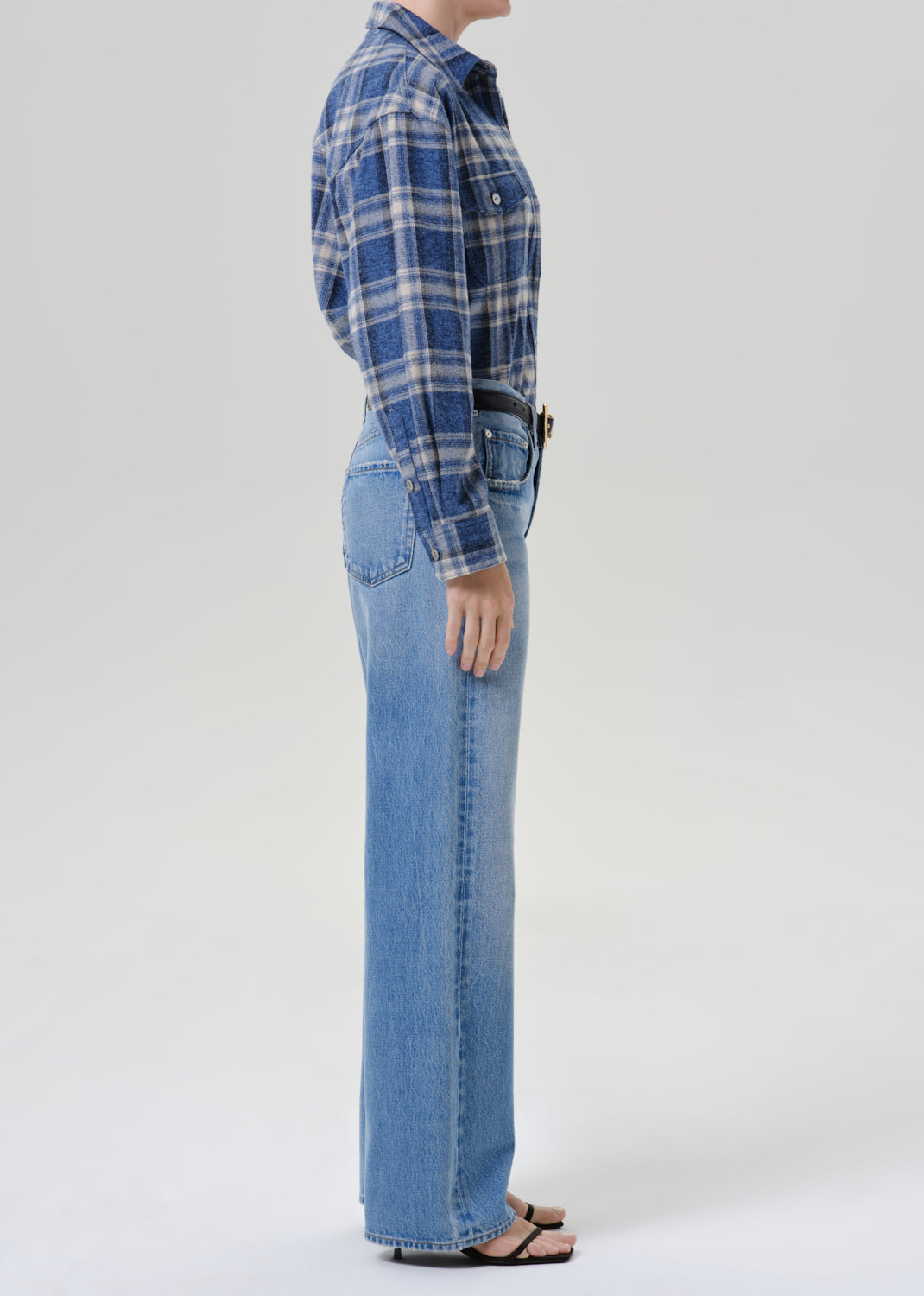 Side profile of a model wearing a blue and white plaid flannel shirt tucked into light-wash blue wide-leg jeans, paired with a black belt and black sandals, against a grey background.
