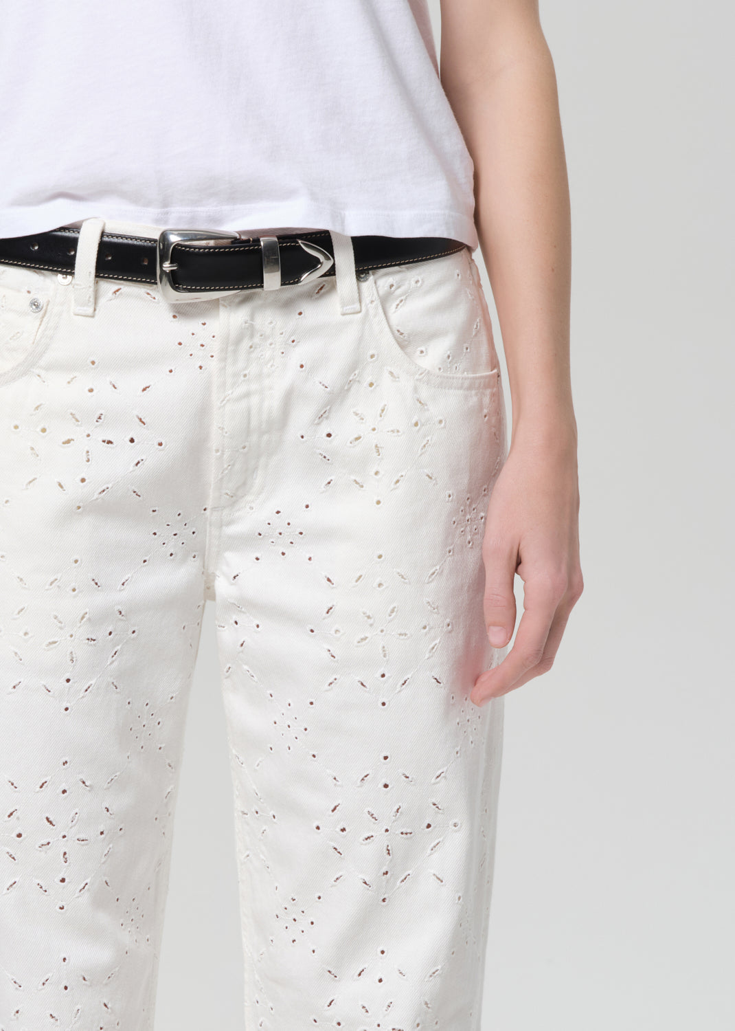Person wearing a white t-shirt and white jeans with eyelet on a plain background