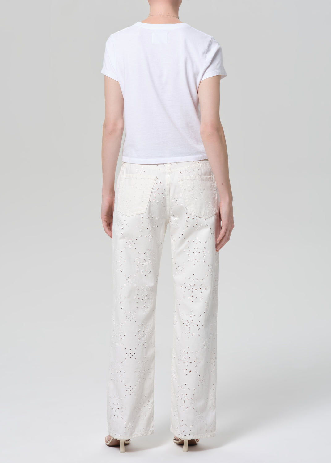 Person wearing a white t-shirt and white jeans with eyelet on a plain background