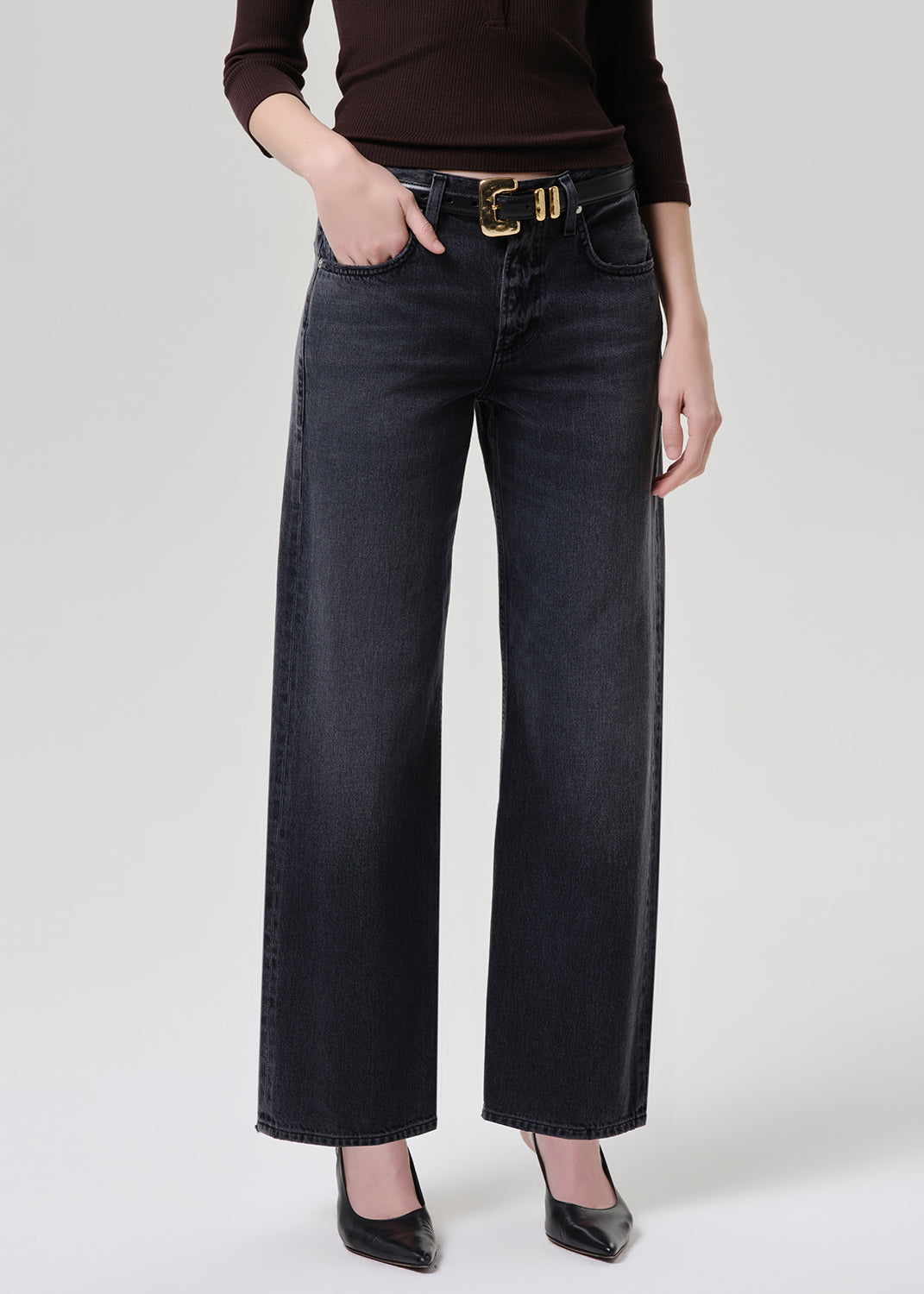 A model wearing dark charcoal wide-leg jeans with a gold-tone belt, paired with a dark brown top and black heels, shown from the waist down on a grey background.