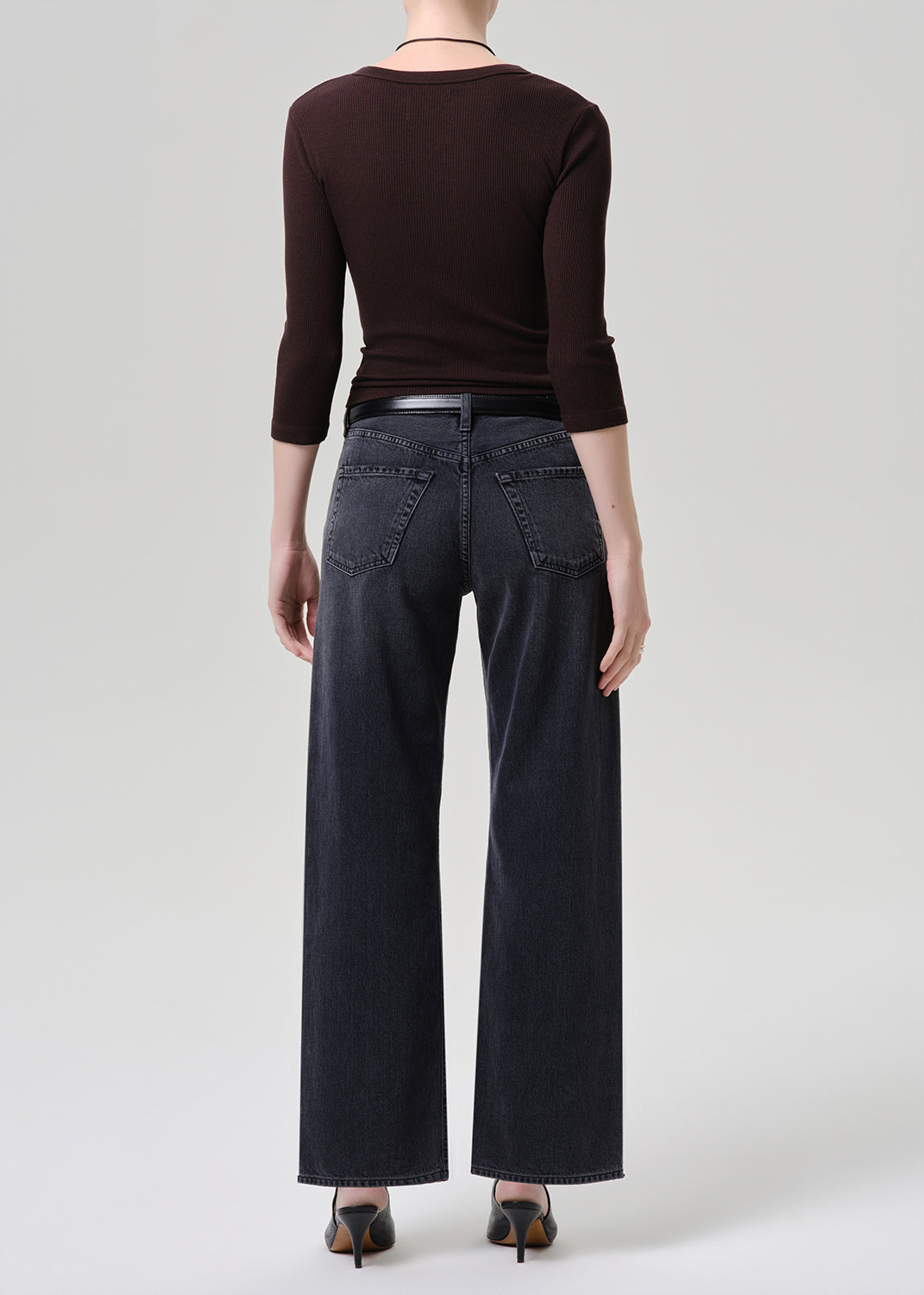 A model shown from the back wearing a dark brown long-sleeve top, dark wide-leg denim jeans, and black heels, set against a grey background.