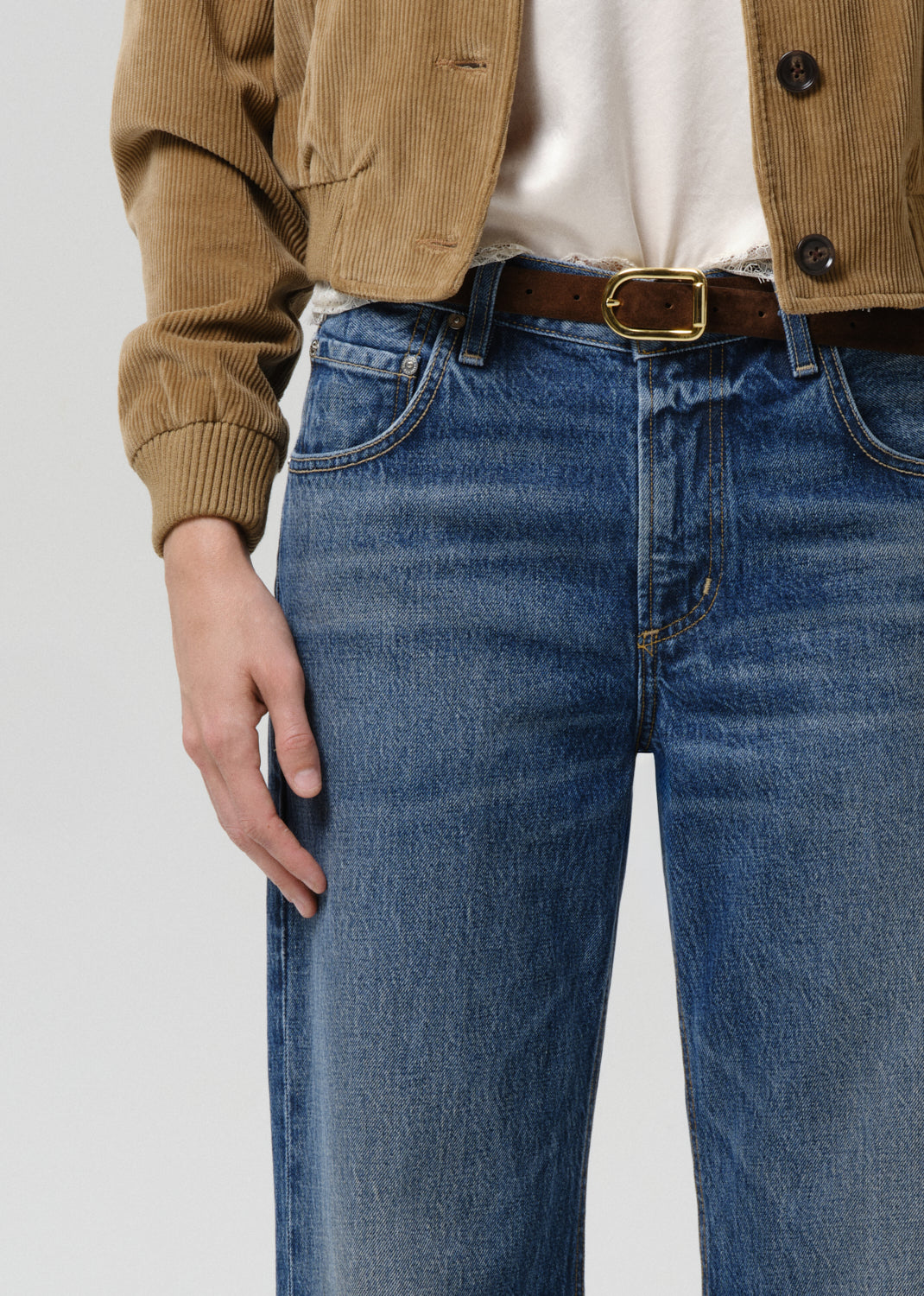 Close-up of a person wearing blue denim jeans with a brown leather belt featuring a gold-tone buckle, styled with a tan corduroy jacket and white top against a grey background.