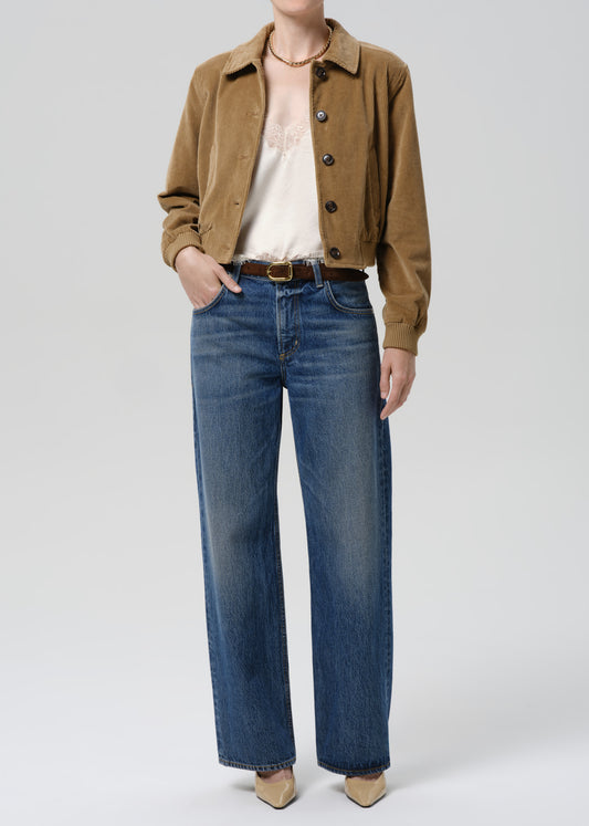 A model wearing a cropped brown corduroy jacket, a white top, and blue wide-leg jeans with a brown belt, standing against a grey background.