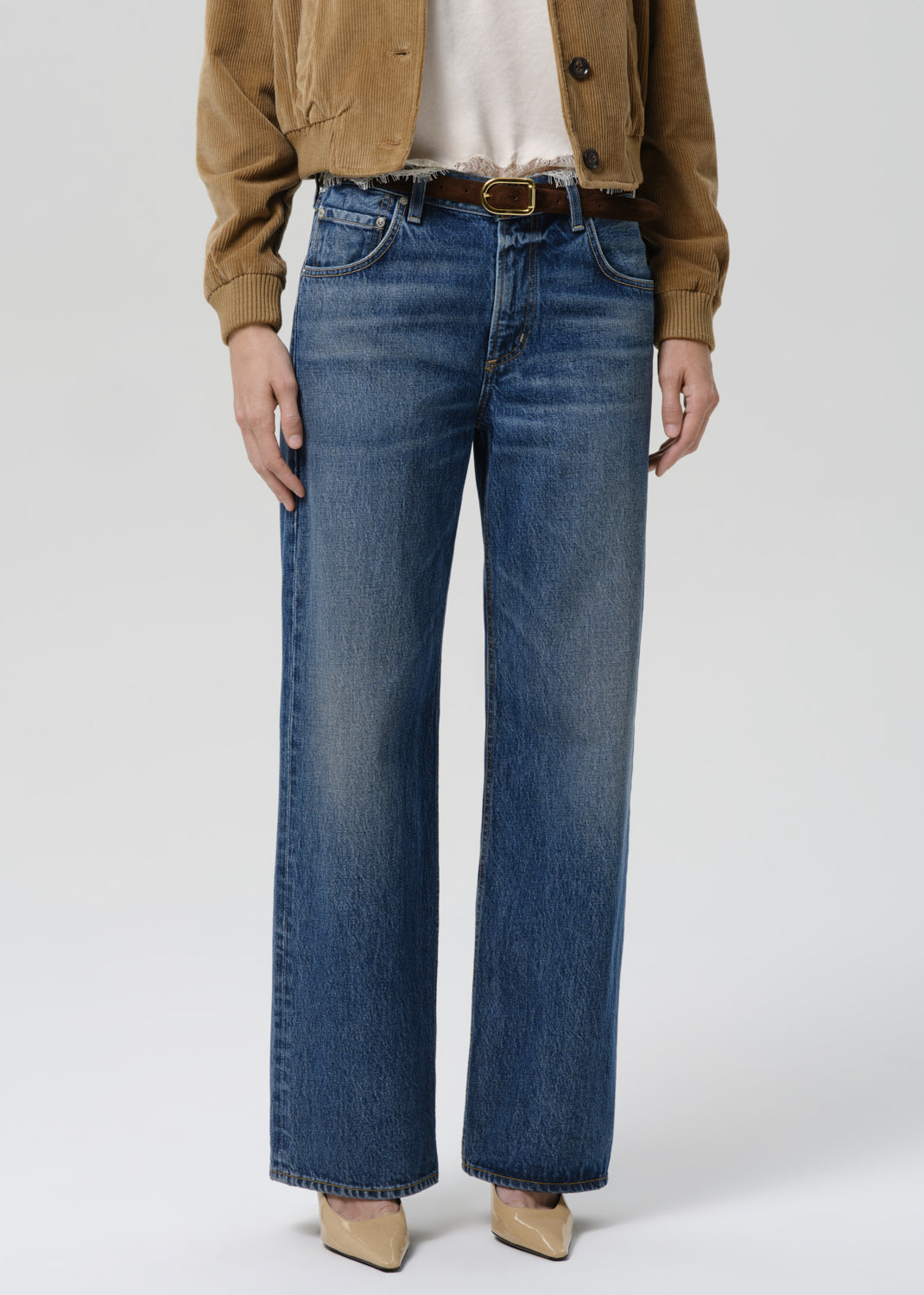 A model wearing a pair of wide-leg blue denim jeans with a brown belt, styled with a tan jacket, white top, and beige heels against a grey background.
