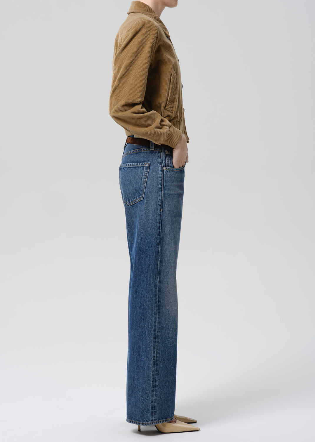 A side-profile view of a model wearing blue straight-leg jeans, a brown jacket, and beige pointed-toe flats against a grey background.