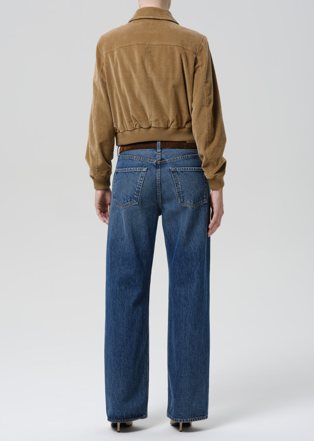 Rear view of a model wearing a cropped tan corduroy jacket and blue wide-leg jeans against a grey background.