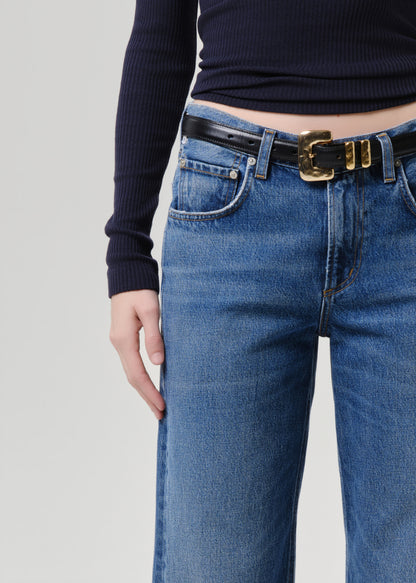 Close-up of a person wearing blue jeans with a black leather belt featuring a prominent gold buckle, styled with a dark navy ribbed long-sleeve top against a grey background.