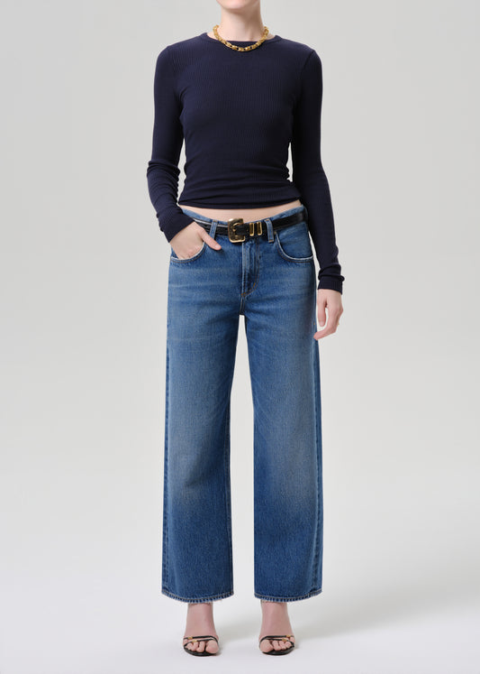 A model wearing a navy blue long-sleeve top, blue wide-leg denim jeans with a gold-tone belt, and black sandals, standing against a grey background.