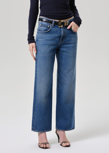 A model wearing a pair of wide-leg blue denim jeans with a black belt, paired with a black long-sleeve top and black heeled sandals, photographed against a grey background.