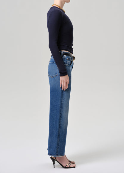 A model shown in profile wearing a navy blue long-sleeve top, high-waisted blue jeans, and black heeled sandals against a grey background.