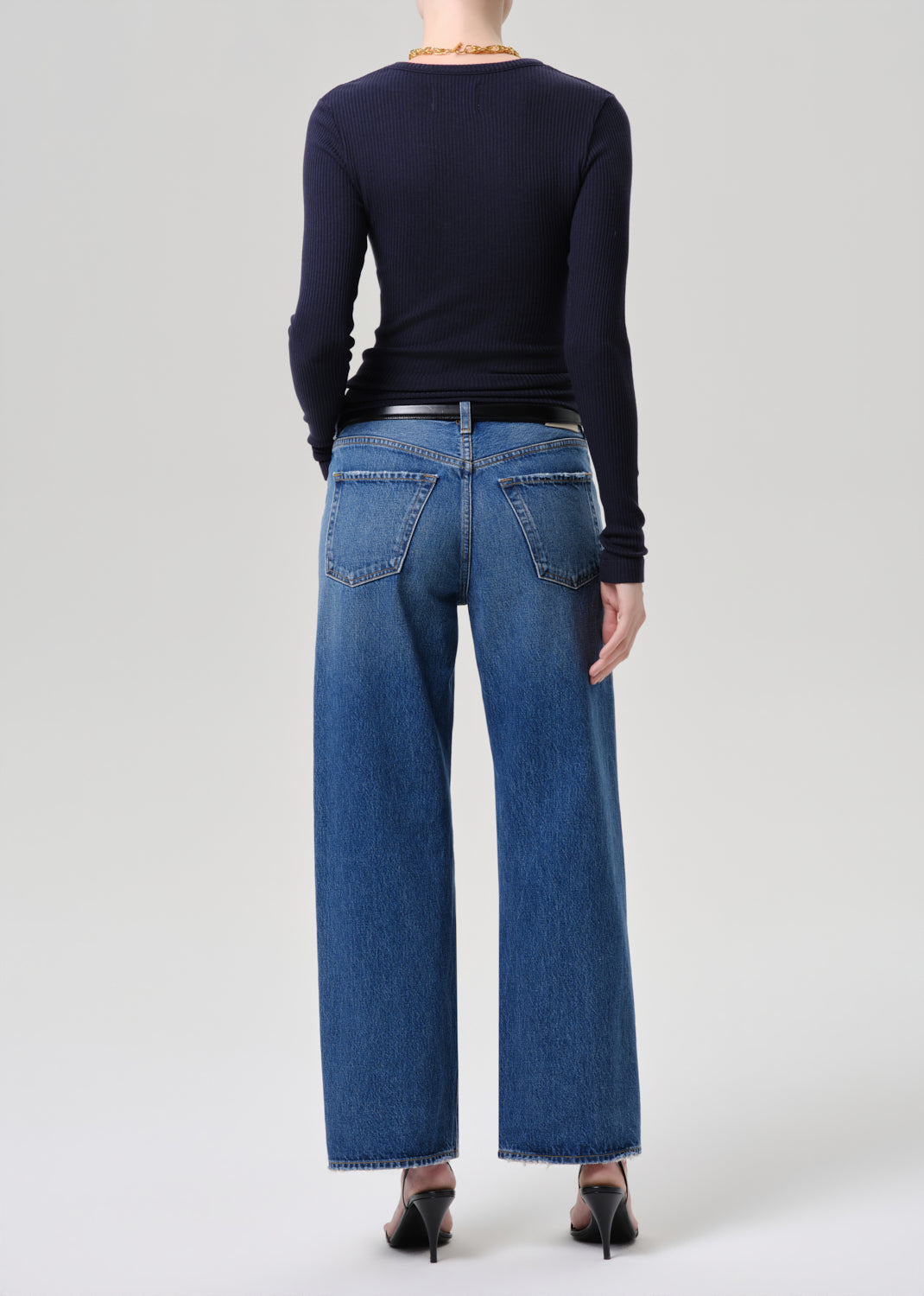A model shown from the back wearing a navy long-sleeve top, blue wide-leg jeans, a black belt, and black heels against a grey background.