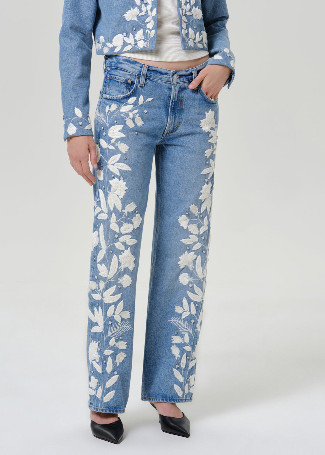 Woman wearing light blue floral jeans with a matching light blue floral denim jacket on a grey background.