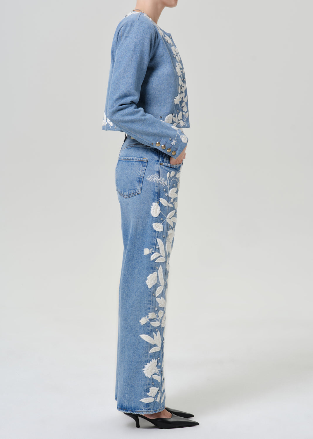 Woman wearing light blue floral jeans with a matching light blue floral denim jacket viewed from the side on a grey background.